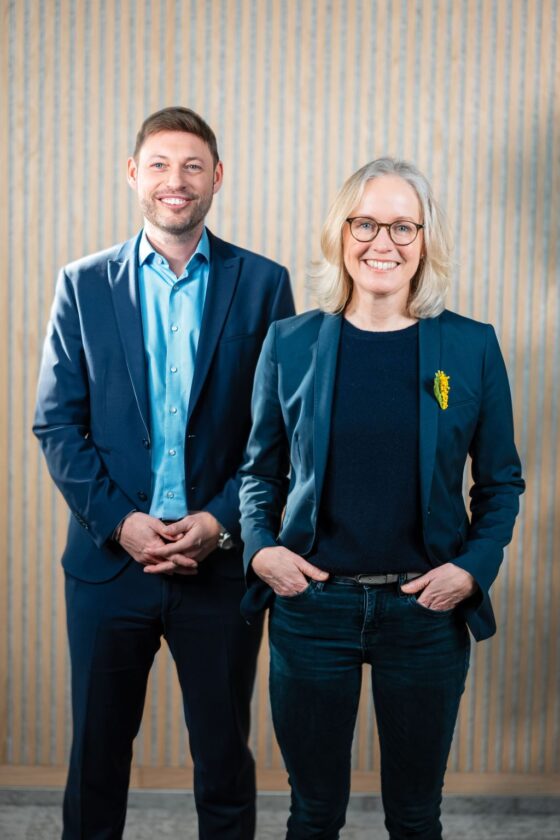 Ein Mann und eine Frau stehen nebeneinander. Der Mann trägt einen blauen Anzug mit einer hellblauen Hemd und hat die Hände vor sich gefaltet. Die Frau trägt eine dunkle Bluse unter einem blauen Blazer und hat ihre Hände in die Hüften gestützt. Auf ihrem Blazer befindet sich eine gelbe Blume. Im Hintergrund sind vertikale Streifen an der Wand zu sehen.