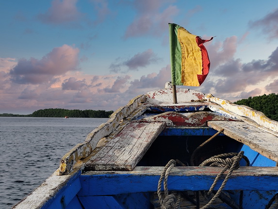 Ein Boot mit einer abgenutzten blauen Oberfläche und einem beschädigten Holzdeck. An der Rückseite weht eine rot-gelb-grüne Flagge. Im Hintergrund erstreckt sich das Wasser unter einem bewölkten Himmel mit sanften Wolken, während eine grüne Küstenlinie sichtbar ist.