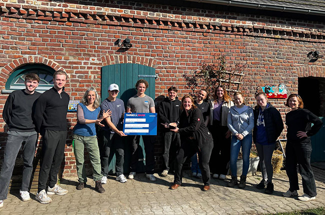 Eine Gruppe von Personen steht vor einer Backsteinmauer mit einer blauen Tür. Sie halten ein Schild in der Hand. Der Hintergrund zeigt Pflanzen und einen Teil eines Schuppens.