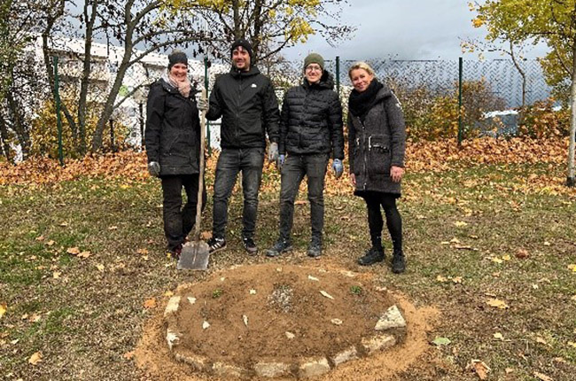 Vier Personen stehen auf einem Rasen mit herbstlichen Blättern. Sie sind in dunkler Kleidung gekleidet und wirken, als ob sie an einem Projekt oder einer Zeremonie beteiligt sind. Vor ihnen liegt ein kreisförmiger Bereich mit Erde und Steinen, möglicherweise ein neu gepflanzter Baum oder eine Gedenkstätte. Im Hintergrund sind Bäume und ein Zaun sichtbar.