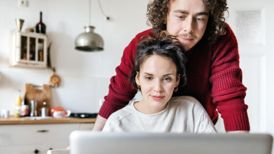Zwei Personen in einer hellen Küche. Eine Person sitzt vor einem Laptop und trägt ein helles Oberteil. Die andere Person steht hinter ihr, trägt einen roten Pullover und hat lockige Haare. Im Hintergrund sind Küchenschränke und verschiedene Kochutensilien sichtbar.