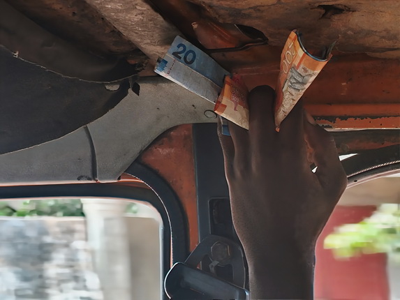 Eine Hand hält Geldscheine, die unter dem Dach eines Fahrzeugs befestigt sind. Ein blauer 20-Schein und ein orangefarbener 100-Schein sind sichtbar.