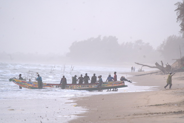 Eine Gruppe von Menschen steht an einem Strand in der Nähe eines farbenfrohen Fischerbootes. Der Himmel ist bewölkt und neblig, während die Wellen sanft gegen den Strand schlagen. Im Hintergrund sind Bäume und einige Personen zu sehen, die am Ufer spazieren.