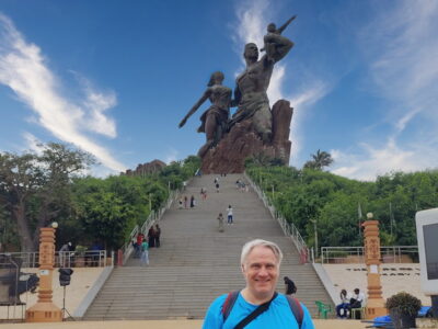 Eine große Statue von zwei Menschen und einem Kind, die auf einer Treppe zu sehen sind. Im Hintergrund sind weitere Besucher und Bäume sichtbar. Der Himmel ist blau mit einigen Wolken.