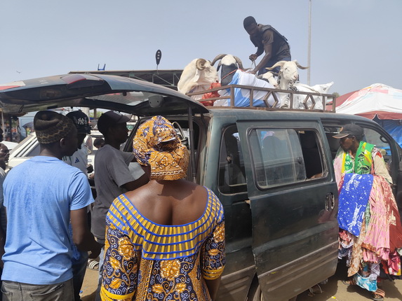 Die Szene zeigt eine Gruppe von Menschen um ein Fahrzeug herum. Auf dem Dach des Fahrzeugs sind zwei Ziegen, während einige Personen mit dem Transport beschäftigt sind. Eine Frau trägt ein buntes Kleid mit einem Muster, während ein Mann in einem blauen T-Shirt daneben steht. Es gibt auch eine andere Person, die auf dem Dach steht, um die Ziegen zu sichern. Der Hintergrund zeigt einen Markt mit Ständen und Zelten.