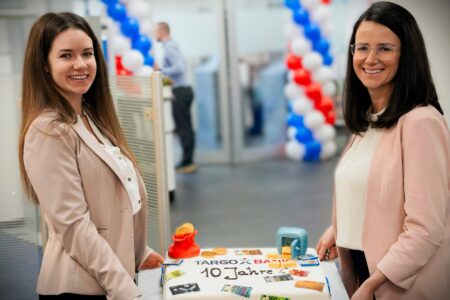 Zwei Frauen stehen neben einer Torte mit der Aufschrift 'TARGO x BANK 10 Jahre'. Die Torte ist dekoriert mit Spielgeld und einer roten Kasse. Im Hintergrund sieht man Luftballons in den Farben Blau, Weiß und Rot.