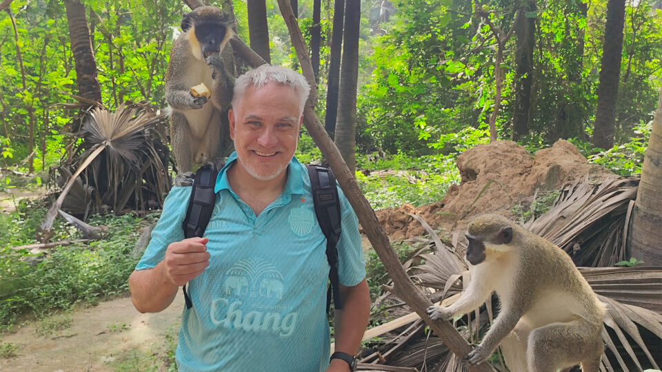 Ein Mann mit grauen Haaren steht in einem Wald, während ein Affe auf seinem Kopf sitzt und ein weiterer Affe auf einem Ast sitzt. Der Mann trägt ein türkisfarbenes Shirt mit einem Logo und hat einen Rucksack auf. Im Hintergrund sind grüne Pflanzen und Bäume sichtbar.