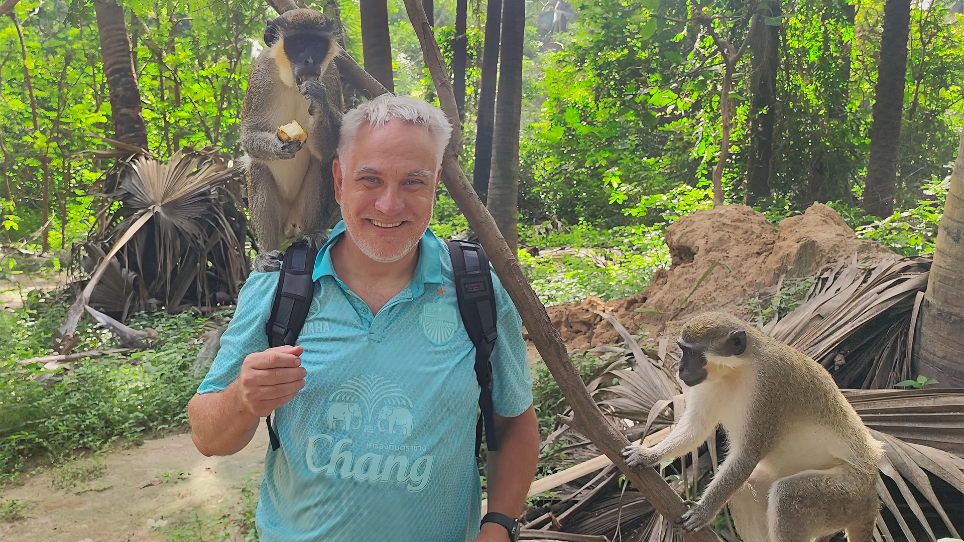 Ein Mann mit grauen Haaren steht in einem Wald, während ein Affe auf seinem Kopf sitzt und ein weiterer Affe auf einem Ast sitzt. Der Mann trägt ein türkisfarbenes Shirt mit einem Logo und hat einen Rucksack auf. Im Hintergrund sind grüne Pflanzen und Bäume sichtbar.