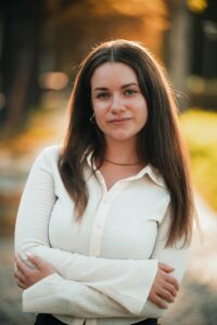 Eine Person mit langen, braunen Haaren steht im Freien. Sie trägt ein helles, langärmliges Hemd und hat die Arme vor der Brust verschränkt. Der Hintergrund zeigt unscharfe Bäume und warmes, goldenes Licht.