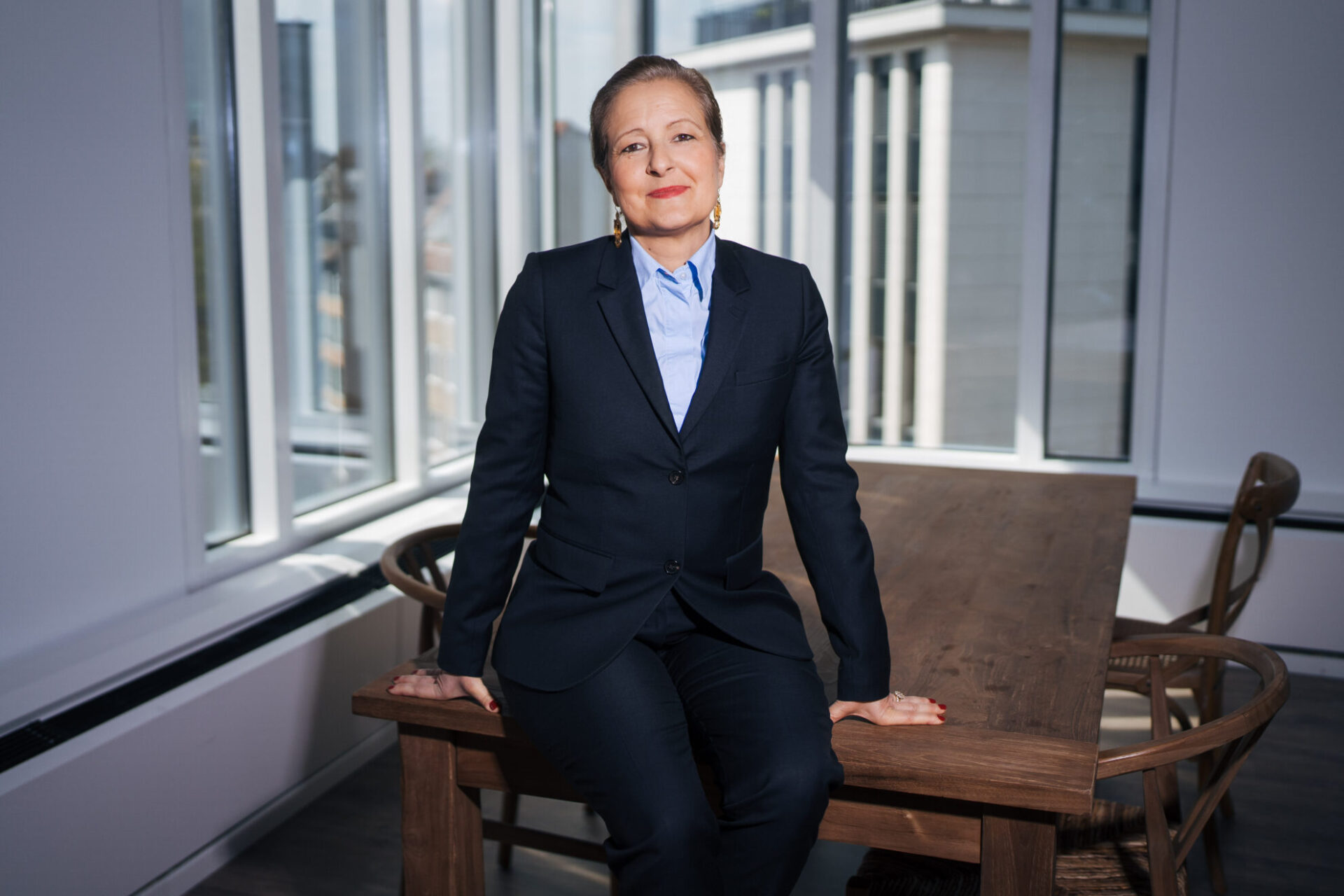 Eine Person in formeller Kleidung sitzt auf einem Tisch in einem modernen Büro mit großen Fenstern. Der Raum hat eine helle, luftige Atmosphäre.