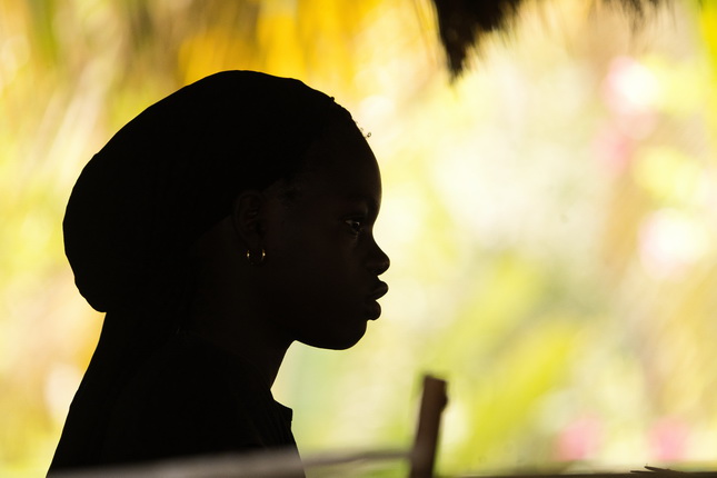 Silhouette einer Person mit einem dunklen Kopftuch, die von einer unscharfen, bunten Hintergrundlandschaft umgeben ist. Die Person ist seitlich dargestellt, und das Gesicht ist nicht sichtbar.