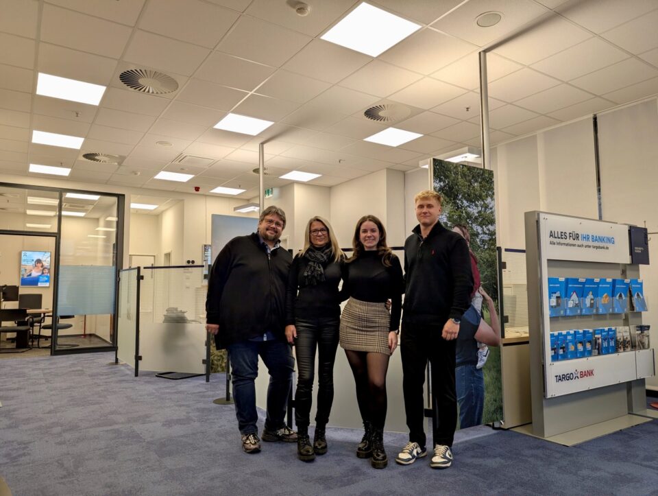 Eine Gruppe von fünf Personen steht in einem modernen Bankbüro. Der Raum hat helle Wände und einen Teppichboden. Im Hintergrund sind Schreibtische und ein Informationsbereich mit Bankmaterialien sichtbar. An der Wand hängt ein Werbebild. Die Personen posieren zusammen, während sie lächeln, und tragen legere Kleidung.