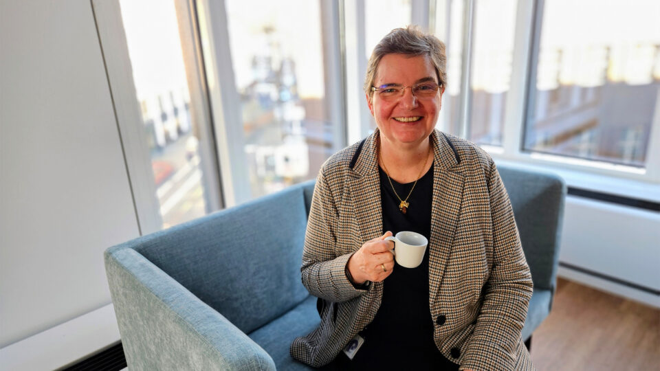 Britta Dahl sitzt auf einem hellblauen Sofa und hält eine kleine Tasse. Die Person trägt eine karierte Jacke und eine schwarze Bluse. Im Hintergrund sind große Fenster, durch die viel Tageslicht strömt. Draußen sind einige Gebäude sichtbar.