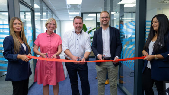 Eine Gruppe von fünf Personen steht in einem modernen Büro und hält ein rotes Band, das sie durchschneiden möchten. Die Personen tragen formelle Kleidung. Im Hintergrund sind Büromöbel und Pflanzen sichtbar.