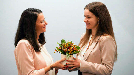 Zwei Frauen stehen gegenüber und halten einen Blumentopf. Eine Frau trägt eine rosa Jacke und die andere eine hellbeige Jacke. Der Blumentopf enthält verschiedene Blumen und grüne Blätter.