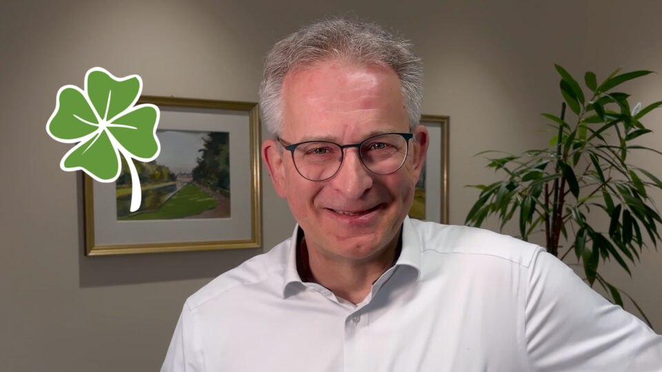 Ein Mann mit grauen Haaren trägt ein weißes Hemd, sein Gesicht ist unscharf. Im Hintergrund befinden sich zwei Bilder in goldenen Rahmen und eine Pflanze.