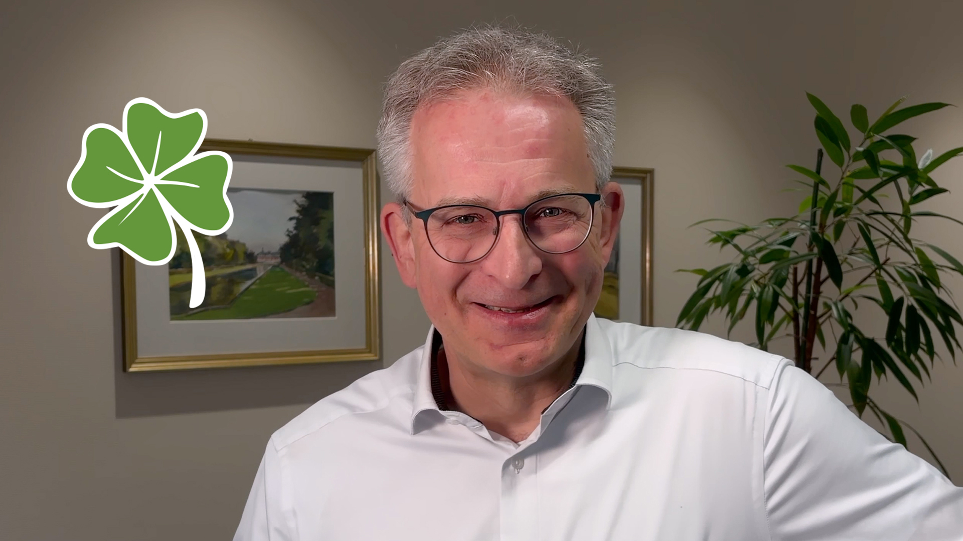 Ein Mann mit grauen Haaren trägt ein weißes Hemd, sein Gesicht ist unscharf. Im Hintergrund befinden sich zwei Bilder in goldenen Rahmen und eine Pflanze.