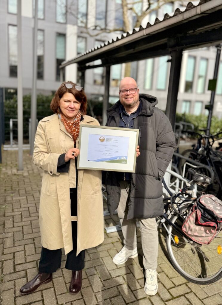 Eine Frau mit braunen Haaren trägt einen beigen Mantel und einen orangefarbenen Schal. Sie hält ein Zertifikat in den Händen. Neben ihr steht ein Mann in einem schwarzen Wintermantel. Im Hintergrund sind Fahrräder und moderne Gebäude zu sehen. Es ist ein sonniger Tag.