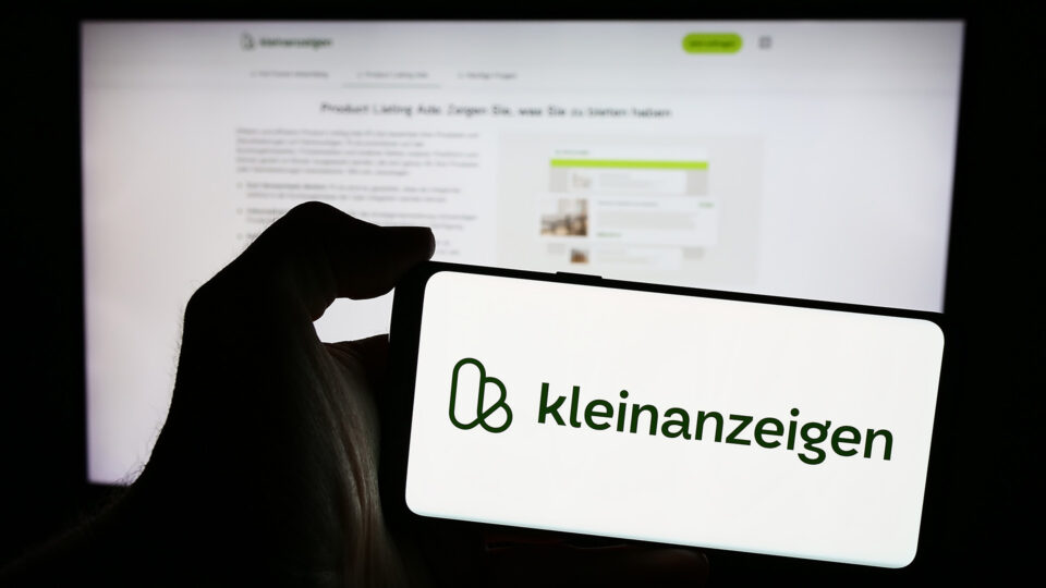Eine Hand hält ein Smartphone, das das Logo von 'kleinanzeigen' zeigt. Im Hintergrund ist ein Computerbildschirm zu sehen, auf dem eine Webseite geöffnet ist, die Informationen über Produkte und Anzeigen zeigt.