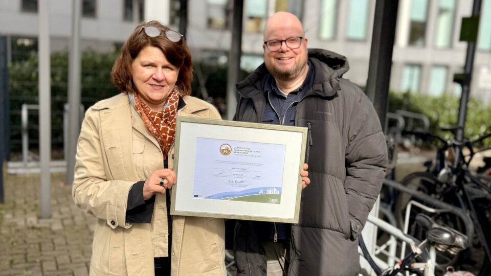 Eine Frau hält ein Zertifikat in der Hand, das einen besonderen Fahrradfreundlichen Standort auszeichnet. Sie trägt eine helle Jacke und ein orangefarbenes Halstuch. Neben ihr steht ein Mann in einer dunklen Jacke. Im Hintergrund sind Fahrräder und moderne Gebäude zu sehen.