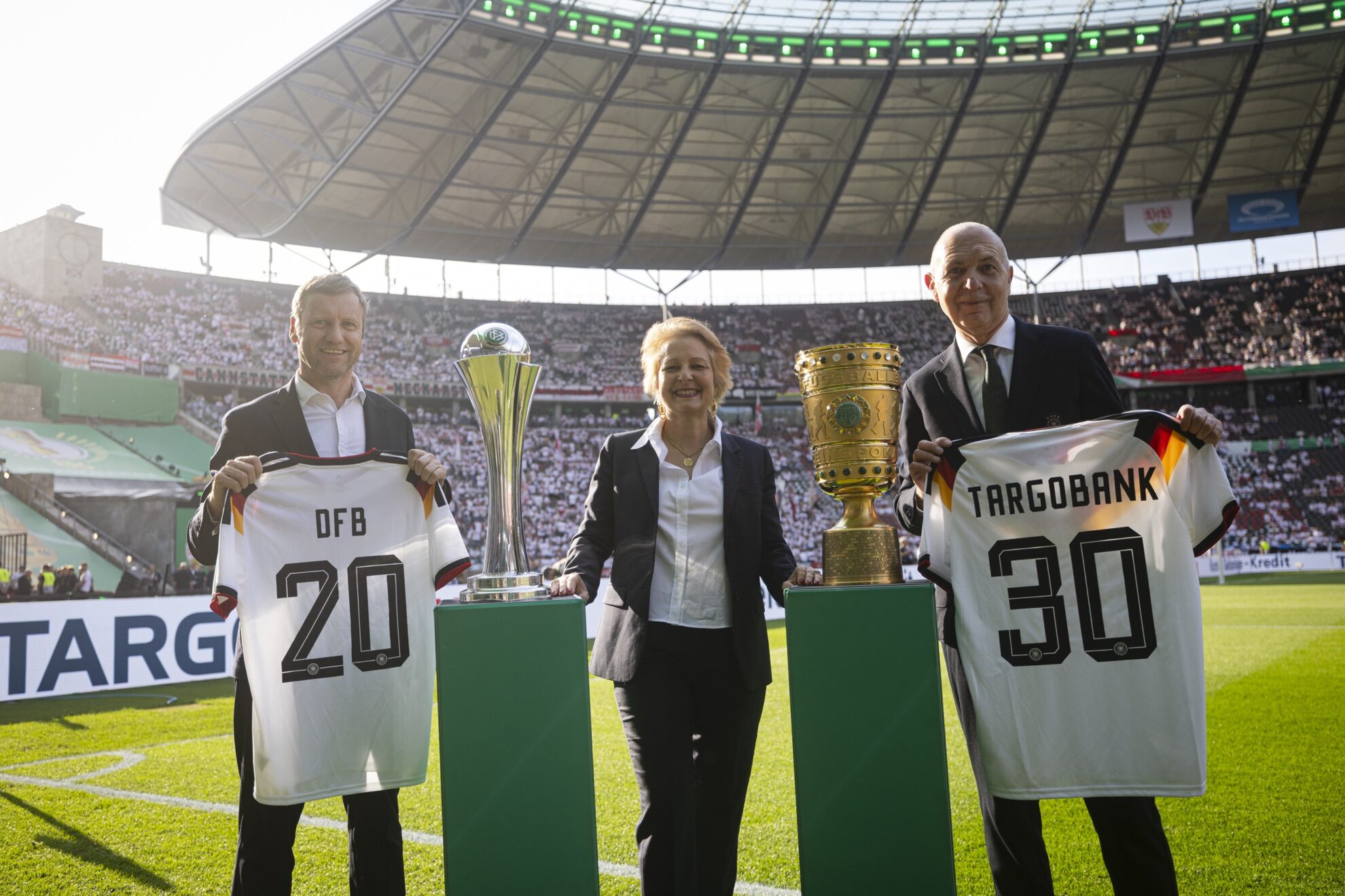 Drei Personen stehen im Stadion. Zwei halten Fußballtrikots mit den Aufschriften 'DFB 20' und 'TARGOBANK 30'. In der Mitte steht eine Frau, die einen Pokal hält. Im Hintergrund sind viele Fans zu sehen.