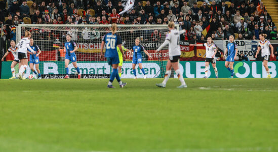 Ein Fußballspiel zwischen zwei Teams, sichtbar sind Spielerinnen in blauen und weißen Trikots. Im Hintergrund sind Fans zu sehen, die Fahnen schwenken. Die Spielerin mit der Nummer 8 hat den Ball, während die Torhüterin in der Mitte steht. Die Zuschauer im Stadion sind aufgeregt.