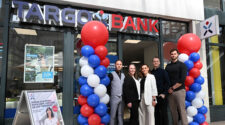 Die TARGOBANK-Filiale mit einer bunten Luftballon-Dekoration in Rot, Blau und Weiß. Vor der Bank stehen fünf Personen, die in formeller Kleidung gekleidet sind. Ein Werbeplakat zeigt den Text 'SO FLEXIBEL, WIE SIE ES WOLLEN' und ein weiteres Plakat hat den Text 'ANDERE SCHLIESSEN, WIR BLEIBEN: WECHSELN SIE ZUR TARGOBANK'.