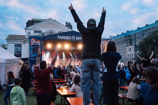 Eine Menschenmenge steht auf einem Festival mit einer Bühne im Hintergrund, auf der Scheinwerfer leuchten. Jemand hebt die Hände in die Luft. Die Atmosphäre ist festlich und lebhaft.