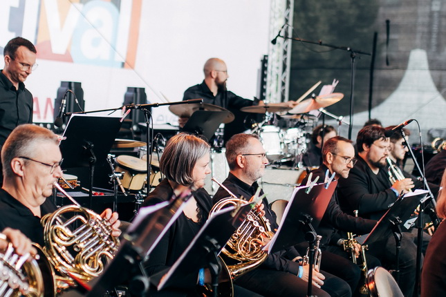 Eine Orchestergruppe, die auf einer Bühne spielt. Musiker mit verschiedenen Instrumenten, darunter Hörner, sitzen in schwarzer Kleidung. Im Hintergrund ist ein Schlagzeug sichtbar. Eine große Leinwand zeigt ein Festival-Logo.