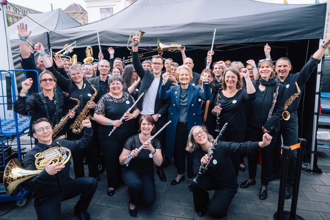 Eine Gruppe von Musikern steht vor einem Zelt und posiert für ein Gruppenfoto. Die meisten tragen schwarze Kleidung und halten Musikinstrumente wie Trompeten, Flöten und Saxophone. Einige heben die Hände in die Luft, während andere lächeln. Im Hintergrund sind weitere Zelte und eine Menschenmenge sichtbar.