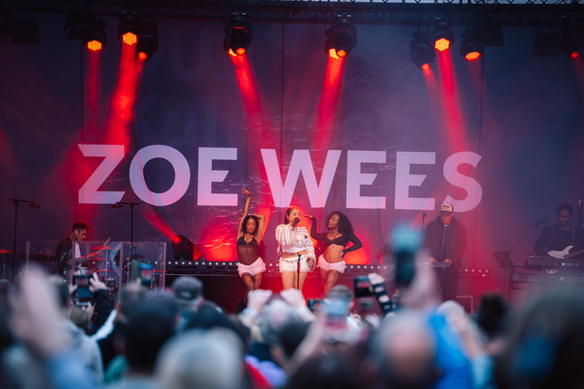 Zoe Wees steht auf einer Bühne und singt, umgeben von Tänzerinnen in schwarz. Im Hintergrund ist das Wort 'ZOE WEES' in großen, weißen Buchstaben zu sehen. Die Bühne ist in rotes Licht getaucht und eine Menschenmenge schaut zu.