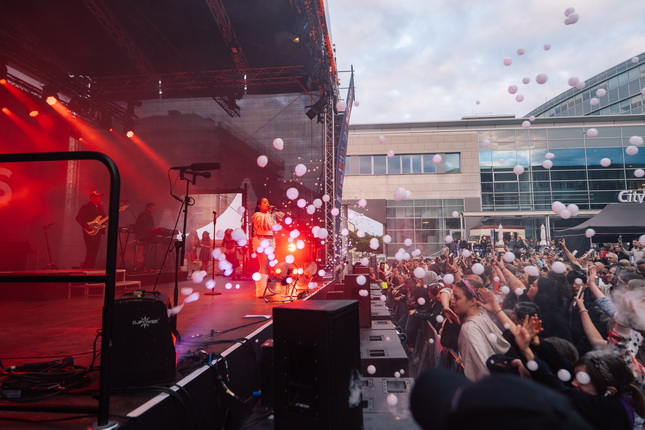 Ein Live-Konzert mit einem Sänger auf der Bühne, umgeben von einer Menge, die mit Luftballons wirft. Die Bühne ist in warmem Licht beleuchtet, während Wolken am Himmel sichtbar sind.