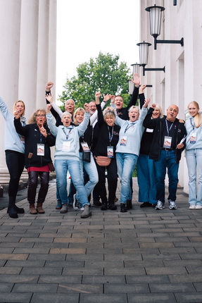 Eine Gruppe von 15 Personen steht auf einem Gehweg, die Hände in die Luft erhoben und lächelt. Sie tragen lässige Kleidung, darunter Jeans und Oberteile in verschiedenen Farben. Im Hintergrund sind Säulen und Laternen sichtbar sowie Bäume. Alle scheinen sich zu freuen und posieren für das Foto.