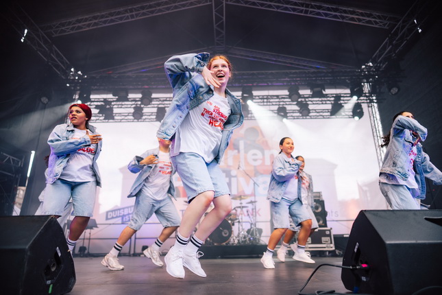 Eine Gruppe von Tänzern führt eine Choreografie auf einer Bühne auf. Sie tragen lässige, hellblaue Jeansjacken und Shorts sowie weiße T-Shirts. Die Tänzer sind in Bewegung und tanzen synchron. Im Hintergrund sind Scheinwerfer und eine große Leinwand sichtbar, die das Event ankündigen. Die Atmosphäre ist energetisch und dynamisch.