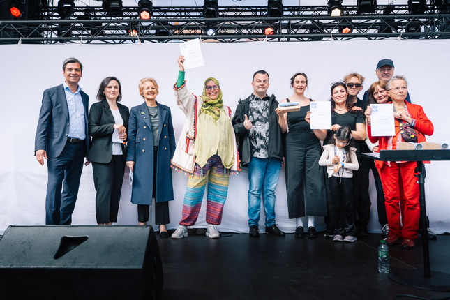Eine Gruppe von Personen steht auf einer Bühne. Einige halten Dokumente in der Hand, darunter eine Person in bunten Kleidern mit einem weißen Dokument. Im Hintergrund sind Lichter und eine weiße Leinwand sichtbar.