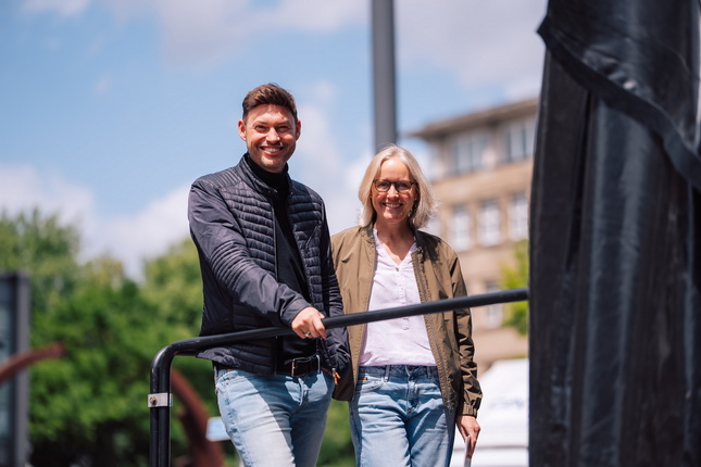 Ein Mann und eine Frau stehen nebeneinander auf einer Plattform. Der Mann trägt eine dunkle Jacke und Jeans, während die Frau eine helle Bluse und eine grüne Jacke trägt. Im Hintergrund sind Bäume und Gebäude sichtbar, und der Himmel ist teilweise bewölkt.