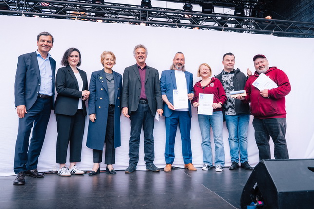 Eine Gruppe von Personen steht auf einer Bühne vor einem weißen Hintergrund. Sie halten Dokumente in den Händen. Die Personen sind unterschiedlich gekleidet und lächeln. Im Vordergrund ist ein Lautsprecher sichtbar.