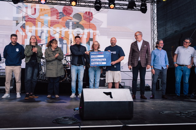 Eine Gruppe von Menschen steht auf einer Bühne, umgeben von Musik-Equipment. Einige applaudieren, während andere auf ein großes Schild schauen, das einen Betrag anzeigt. Im Hintergrund sind bunte Grafiken und ein Musikinstrument zu sehen.