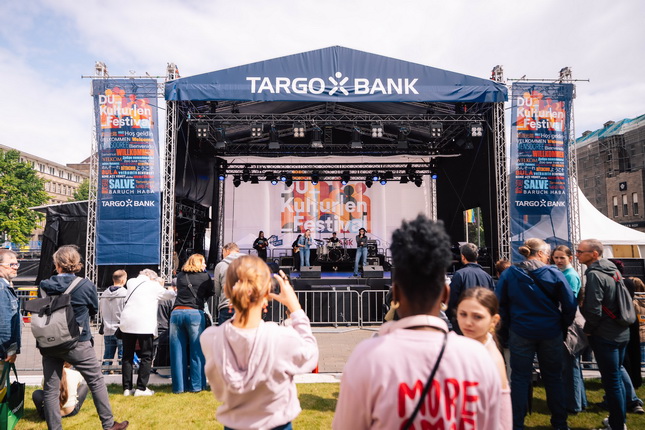 Das Bild zeigt eine Live-Bühne mit dem Banner "DU Kulturen Festival" im Hintergrund. Vor der Bühne stehen Menschen, die auf die Darbietung schauen. Im Vordergrund ist eine Person mit einem weißen Pullover, auf dem "MORE" steht. Die Szene findet bei Tageslicht statt, mit einer Vielzahl von Zuschauern und einem überdachten Bereich hinter der Bühne.