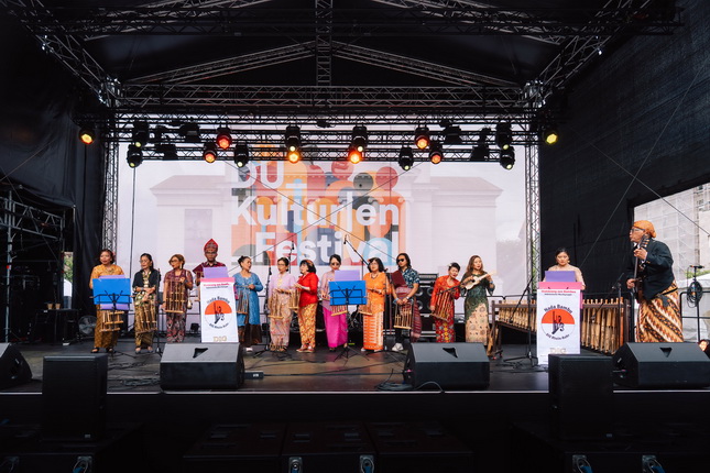 Eine Gruppe von Frauen in traditionellen Kleidern steht auf einer Bühne bei einem Kulturfest. Im Hintergrund sind Scheinwerfer und eine große Leinwand mit dem Schriftzug "Kulturen Fest" zu sehen. Einige Frauen halten Notenständer, während eine Frau spricht. Die Atmosphäre ist festlich und lebhaft.