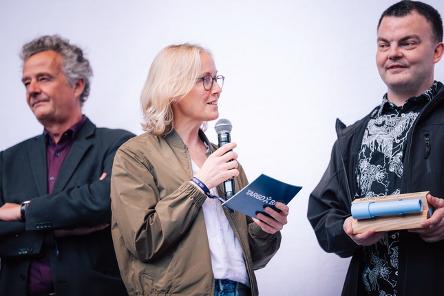 Eine Person mit blonden Haaren hält ein Mikrofon und spricht, während sie ein blaues Blatt Papier in der Hand hält. Neben ihr stehen zwei weitere Personen, eine in einem schwarzen Anzug und die andere in einem dekorierten Hemd. Der Hintergrund ist hell und neutral.