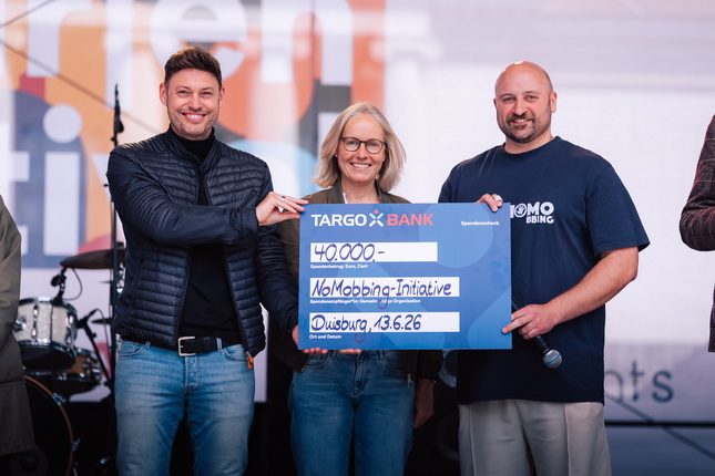 Drei Personen stehen auf einer Bühne und halten einen großen Scheck hoch. Der Scheck ist von TARGO BANK und zeigt einen Betrag von 40.000 Euro für die No-Mobbing-Initiative. Im Hintergrund sind Bühnenlichter und eine unklare Menschenmenge sichtbar.