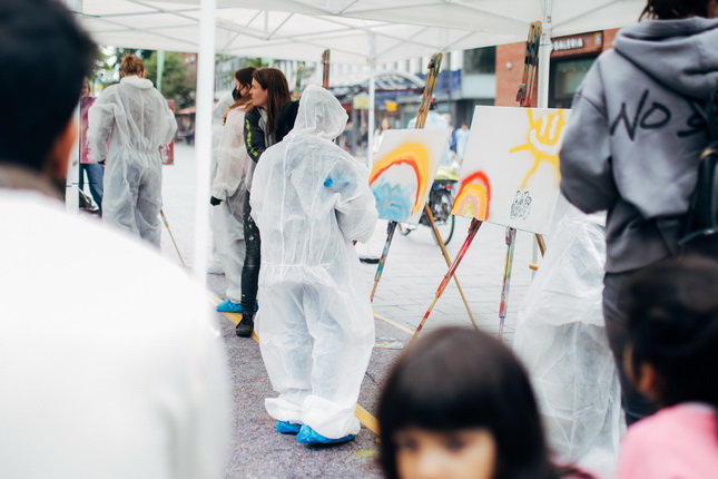 Auf dem Bild sind mehrere Personen sichtbar, die in einem Freiluftbereich unter einem Zelt stehen. Einige tragen Schutzanzüge und arbeiten an Kunstwerken auf Staffeleien. Eine Person steht seitlich mit einem Pinsel in der Hand und malt eine bunte Sonne und einen Regenbogen. Im Hintergrund sind weitere Menschen und Fahrräder zu sehen, während junge Zuschauer auf dem Boden sitzen.