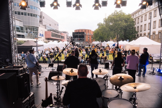 Ein Blick von der Bühne auf ein Musikfestival. Eine Band spielt, während das Publikum im Freien vor ihnen sitzt. Zelte sind im Hintergrund zu sehen, und viele Menschen sind in entspannter Atmosphäre versammelt.