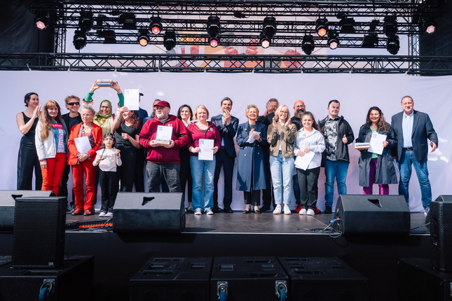Eine Gruppe von Menschen steht auf einer Bühne, lächelt und hält Dokumente in der Hand. Einige Personen tragen farbenfrohe Kleidung, während andere formelle Outfits tragen. Im Hintergrund sind Scheinwerfer und eine große Leinwand erkennbar. Die Szene wirkt festlich und fröhlich.