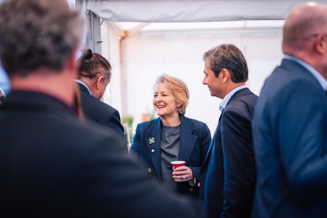 Eine Gruppe von Personen steht in einem Zelt. Eine Frau mit blonden Haaren trägt einen Blazer und hält einen roten Becher. Sie spricht mit mehreren Männern, deren Gesichter unscharf sind. Im Hintergrund sind einige Pflanzen sichtbar.