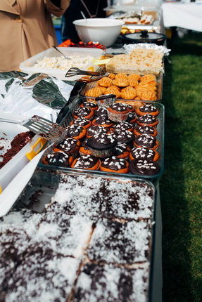 Ein Tisch mit einer Vielzahl von Desserts im Freien. Es gibt Schokoladenkekse, Cupcakes mit Zuckerguss und Kekse. Im Hintergrund sind weitere Speisen zu sehen.