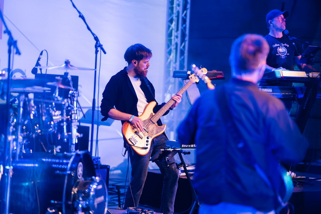 Eine Band spielt live auf einer Bühne. Ein Bassist mit einem gelben Bass steht im Vordergrund, während ein Keyboarder und weitere Musiker im Hintergrund zu sehen sind. Die Beleuchtung ist blau und schafft eine lebhafte Atmosphäre.