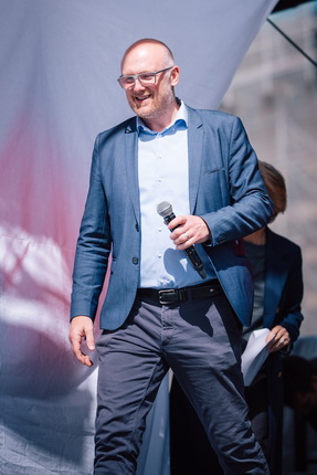 Ein Mann steht auf einer Bühne und hält ein Mikrofon in der Hand. Er trägt einen blauen Anzug und ein hellblaues Hemd. Im Hintergrund ist eine Person zu sehen, die nicht klar identifizierbar ist.