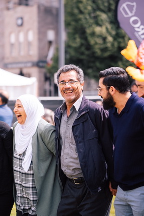 Die Abbildung zeigt mehrere Personen, die nebeneinander stehen. Eine Person trägt ein Kopftuch, während eine andere eine Jacke und ein Hemd trägt. Die Umgebung scheint eine Veranstaltung im Freien zu sein, mit einer unklaren Hintergrundszene.